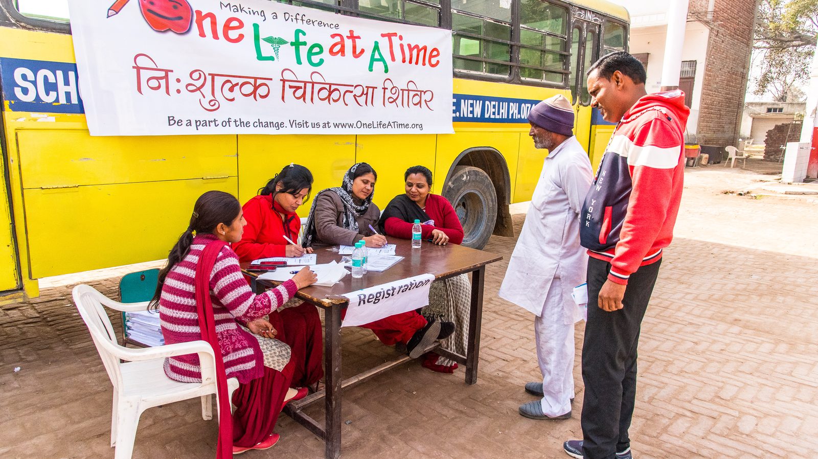 Volunteers at a One Life At A Time medical-camp registration desk beside a school bus